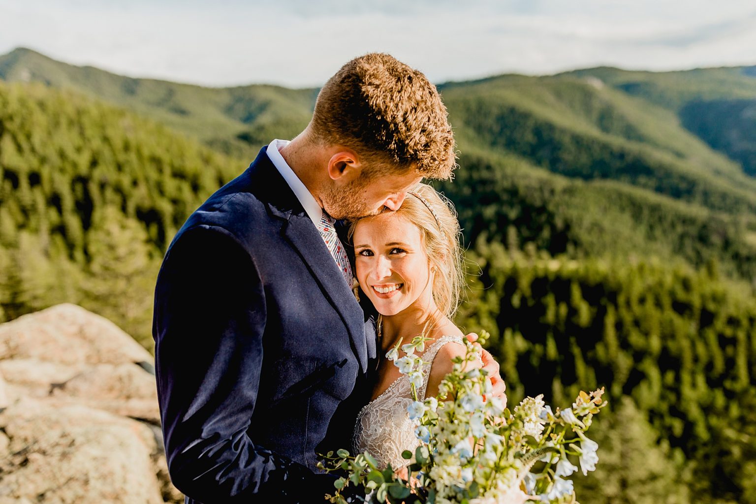 groom kisses bride on the forehead with beautiful mountain views and bouquet showing