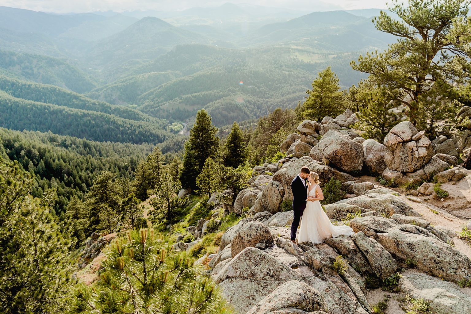 couple stands on the rocks hugging one another with beautiful green mountains, trees, and rocks in the background of the Rocky Mountains in colorado