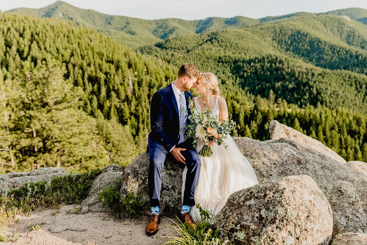 couple sit together on a rock with green tree mountain background at sunset of colorado