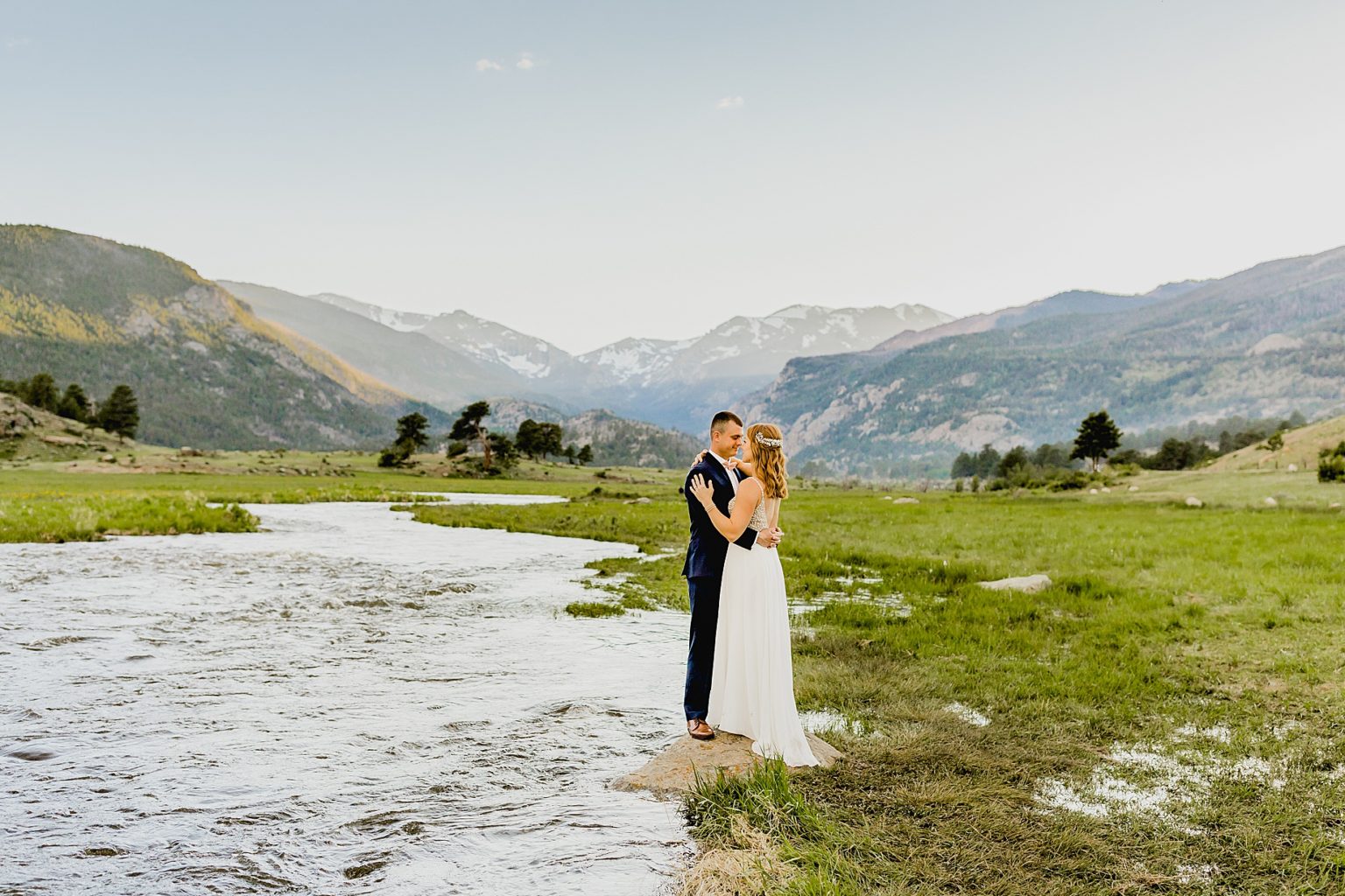 bride and groom stand on rock next to flowing colorado river with mountain background