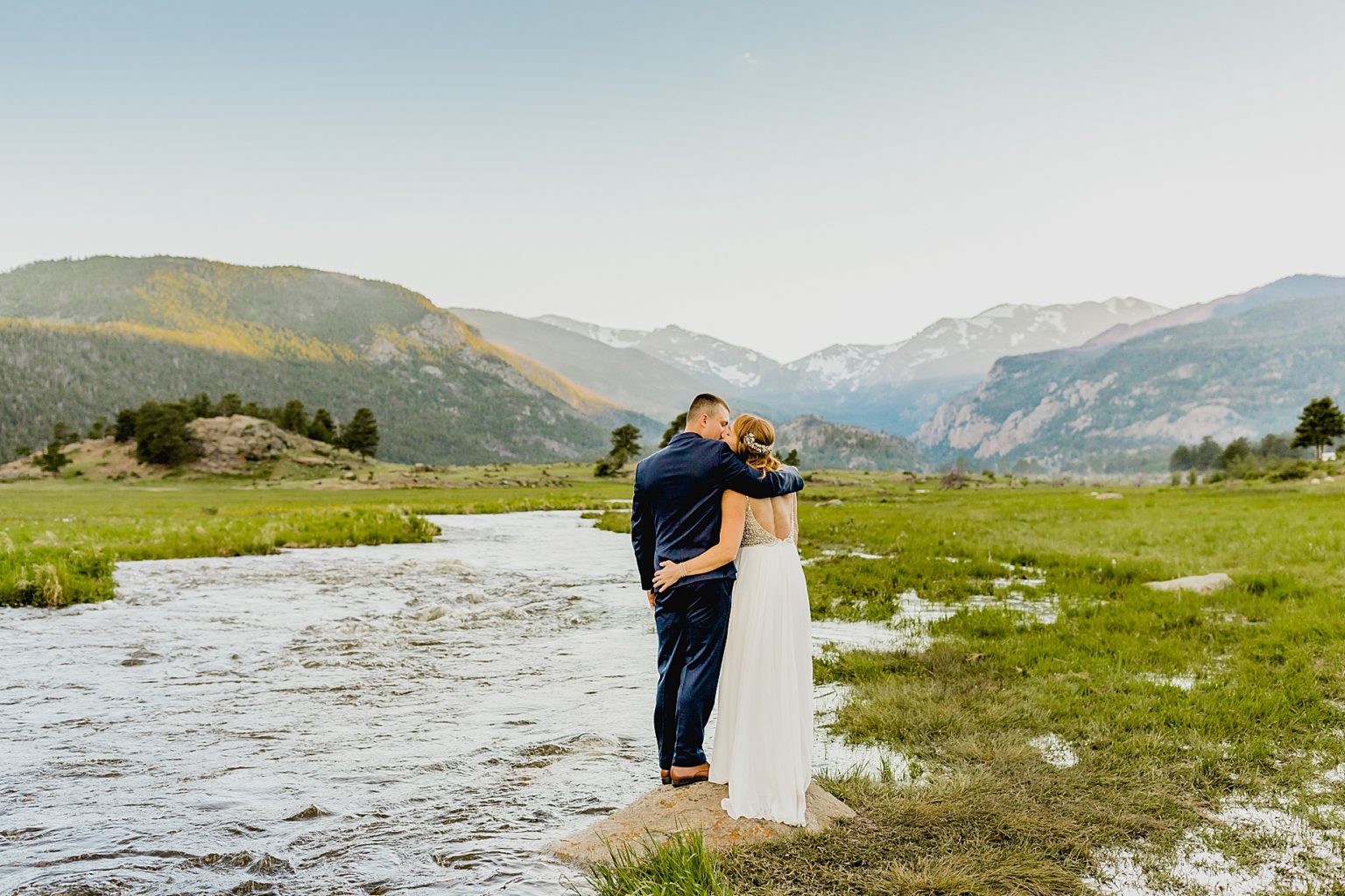 bride and groom stand on rock next to flowing colorado river with mountain background