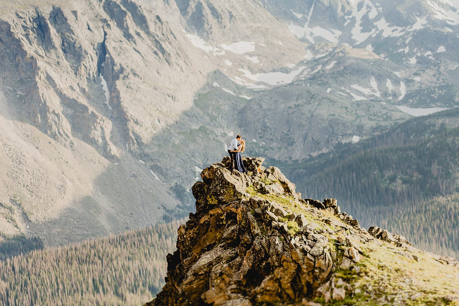 couple embraces on Rocky Mountain cliff edge with gorgeous mountain scenery in the background