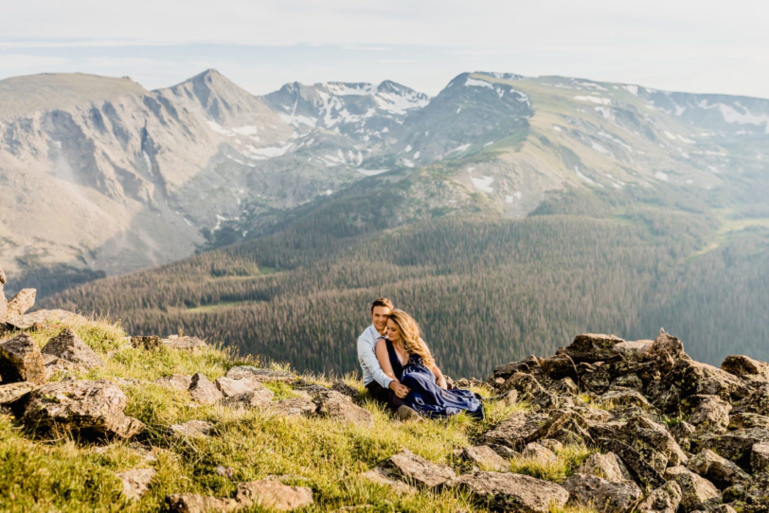couple sits in the wildflowers and grass while embracing and smiling with stunning Rocky Mountain background