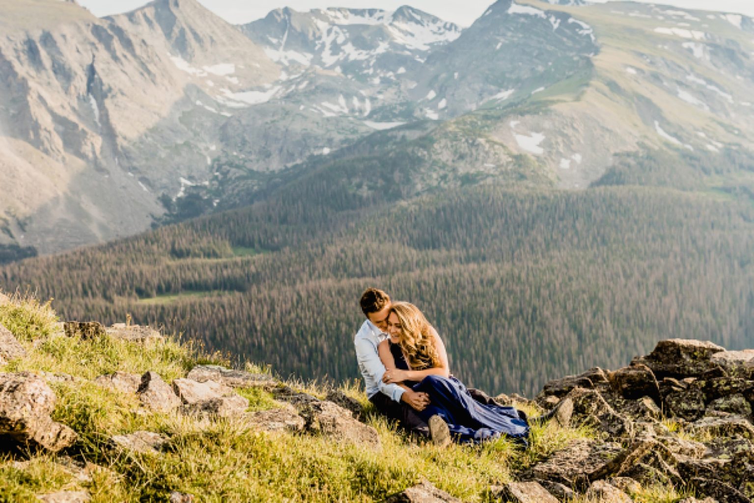couple sits in the wildflowers and grass while embracing and smiling with stunning Rocky Mountain background