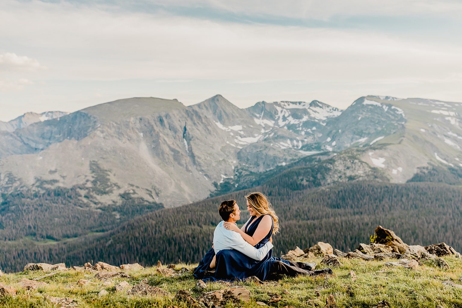 man and woman sit in the grass overlooking mountain backdrop with beautiful colors