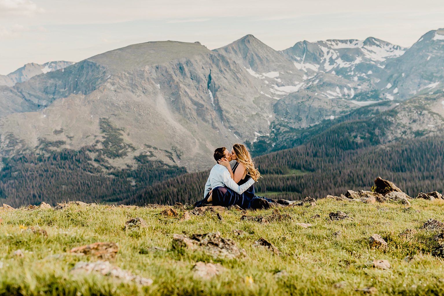 man and woman sit in the grass overlooking mountain backdrop with beautiful colors