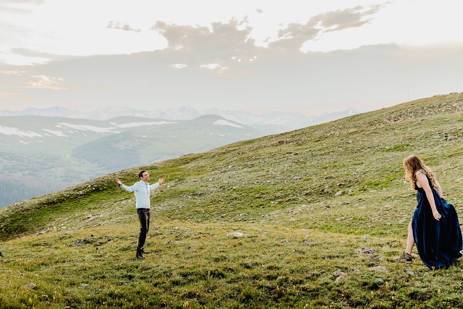woman gets ready to run into man's arms for a big hug in front of the stunning Rocky Mountains in the green summer grass