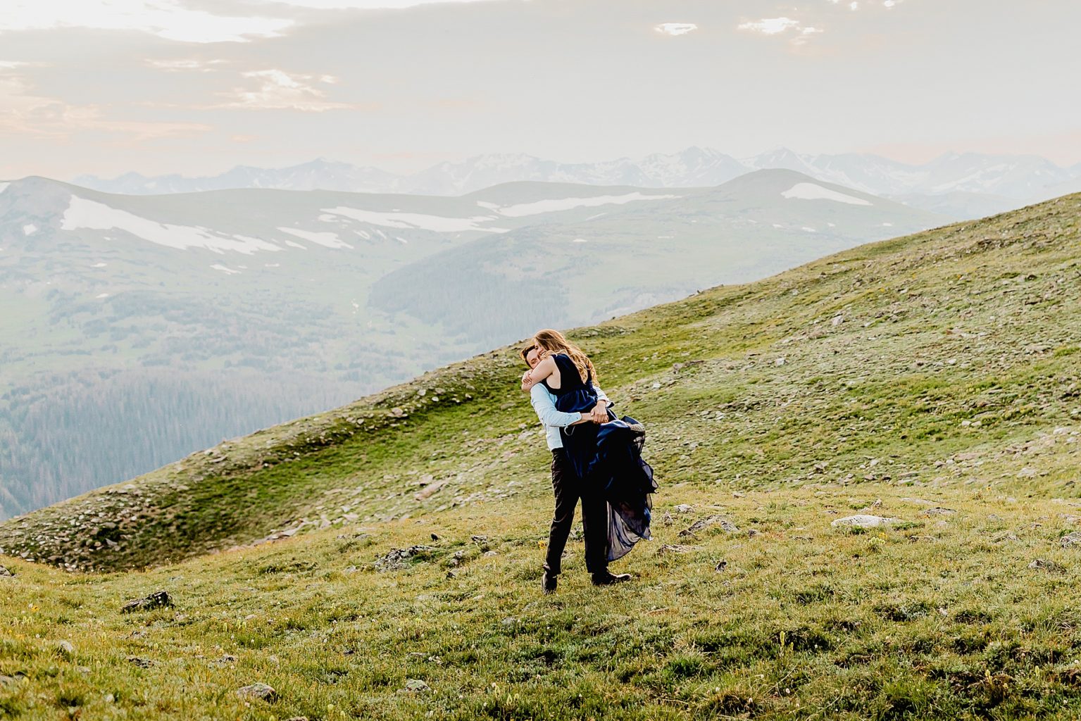 man lifts woman in front of the stunning Rocky Mountains in the green summer grass