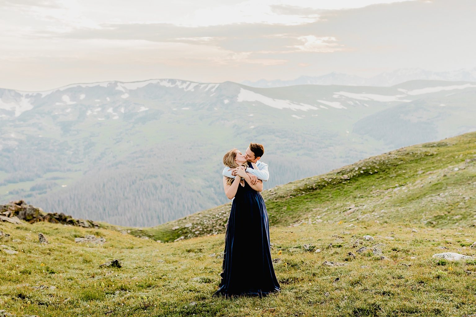 man hugs woman in front of the stunning Rocky Mountains in the green summer grass