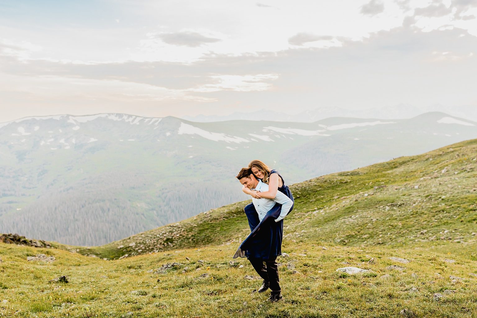 man gives woman piggyback ride in the green summer grass with mountain backdrop