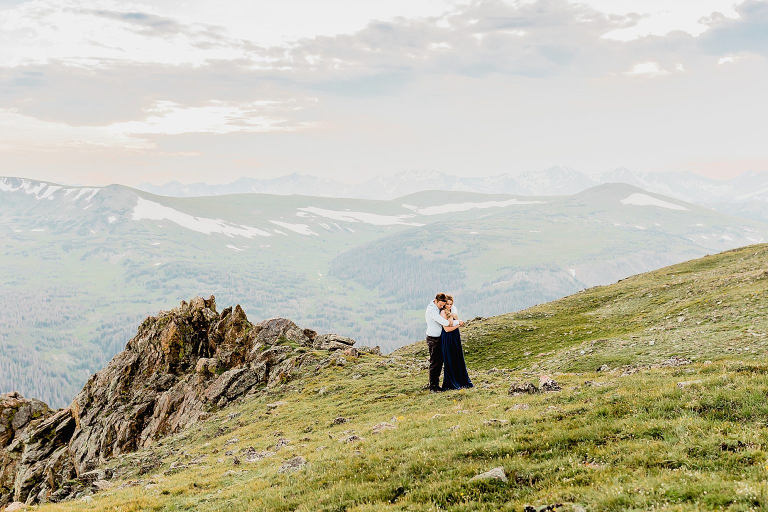 man hugs woman in front of the stunning Rocky Mountains in the green summer grass