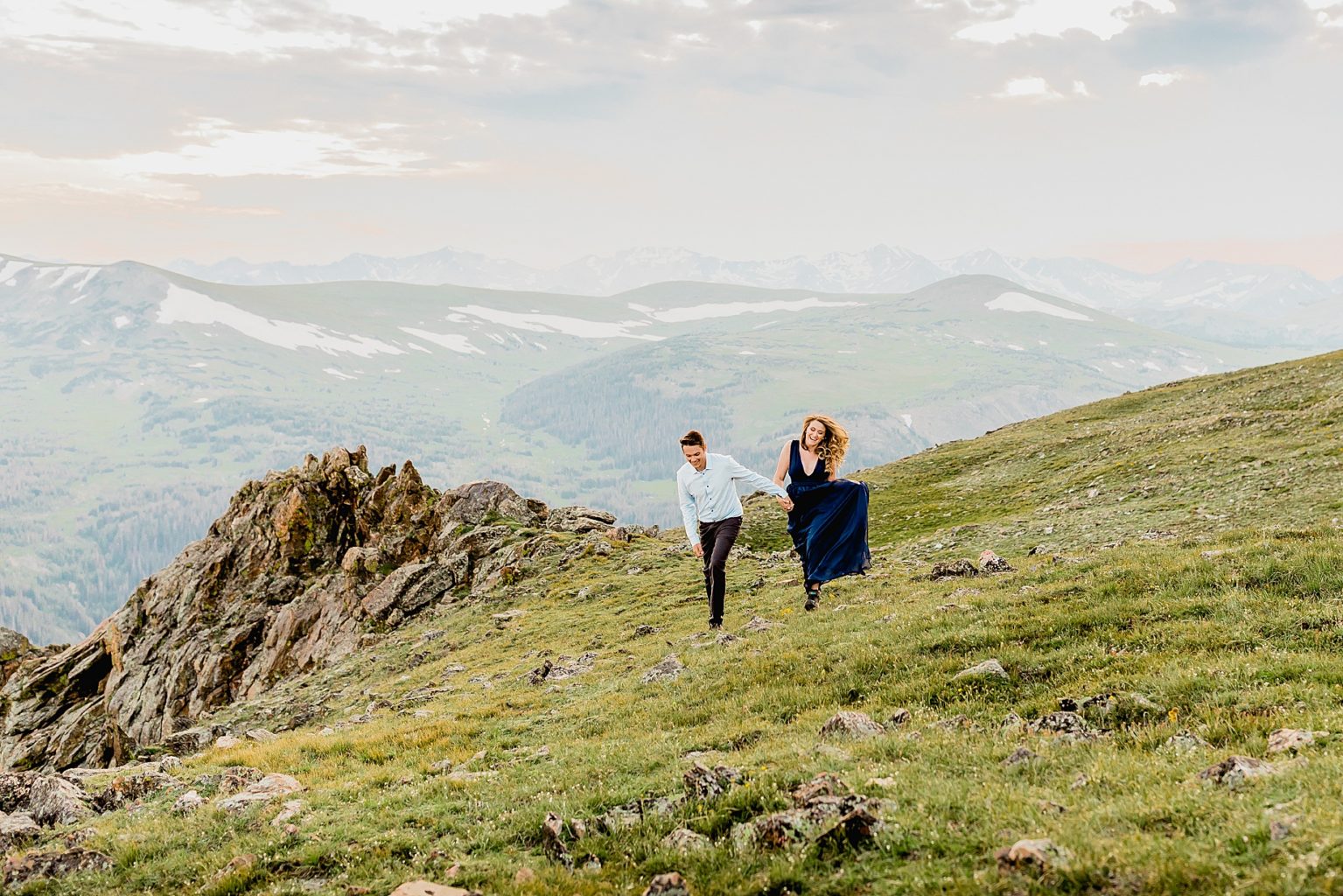 man and woman skip holding hands while laughing in the green grass with majestic Rocky Mountain scenery