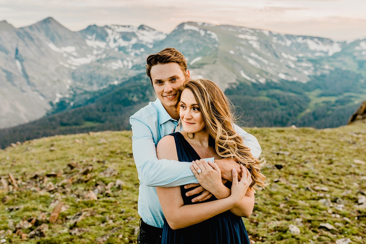 man gives woman a hug and kiss with majestic rocky mountain and green grass scenery in the background