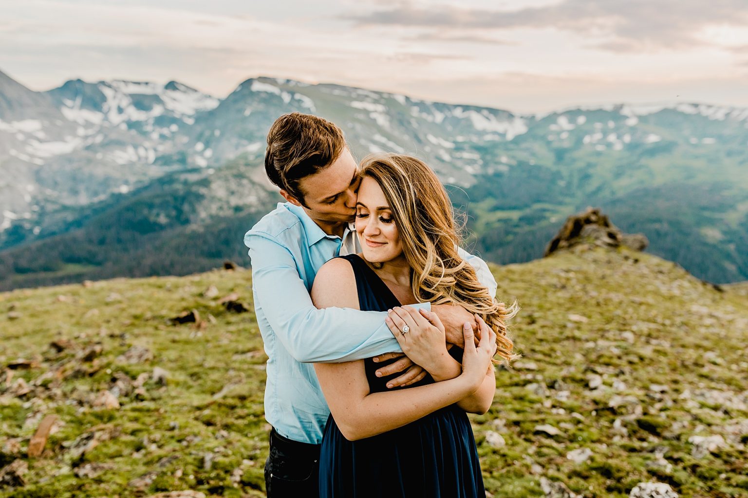 man gives woman a hug and kiss with majestic rocky mountain and green grass scenery in the background