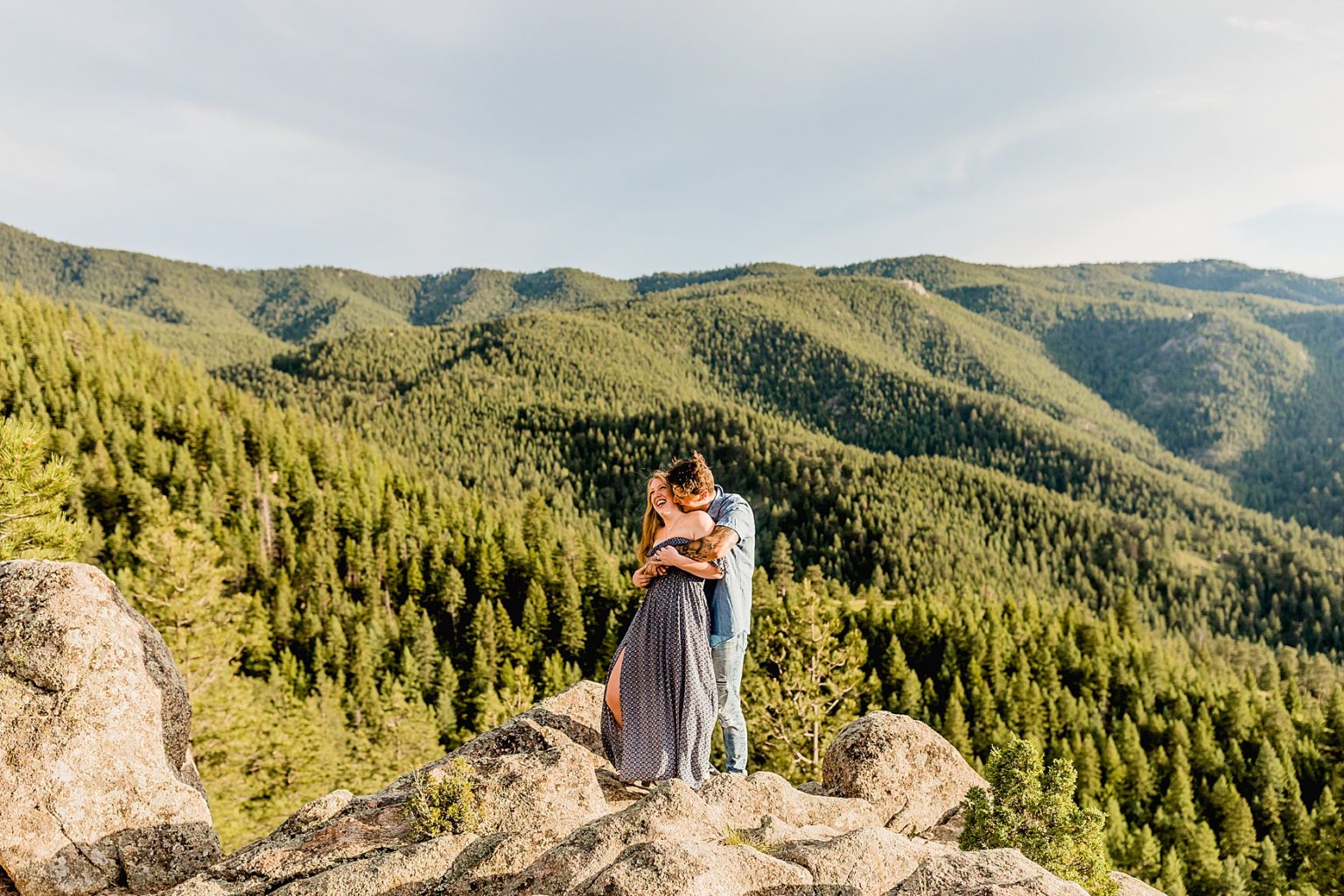 man hugs woman from behind with laughter in a gorgeous green mountain and evergreen tree setting