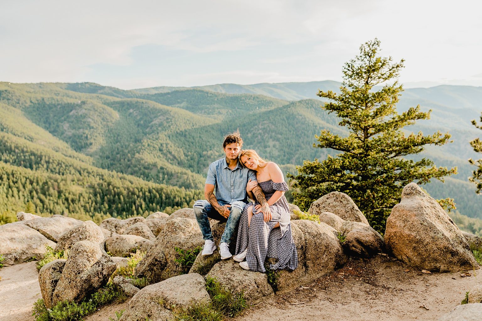 couple sits on a rock holding each other with green mountains and trees in the distance showing off boulder colorado