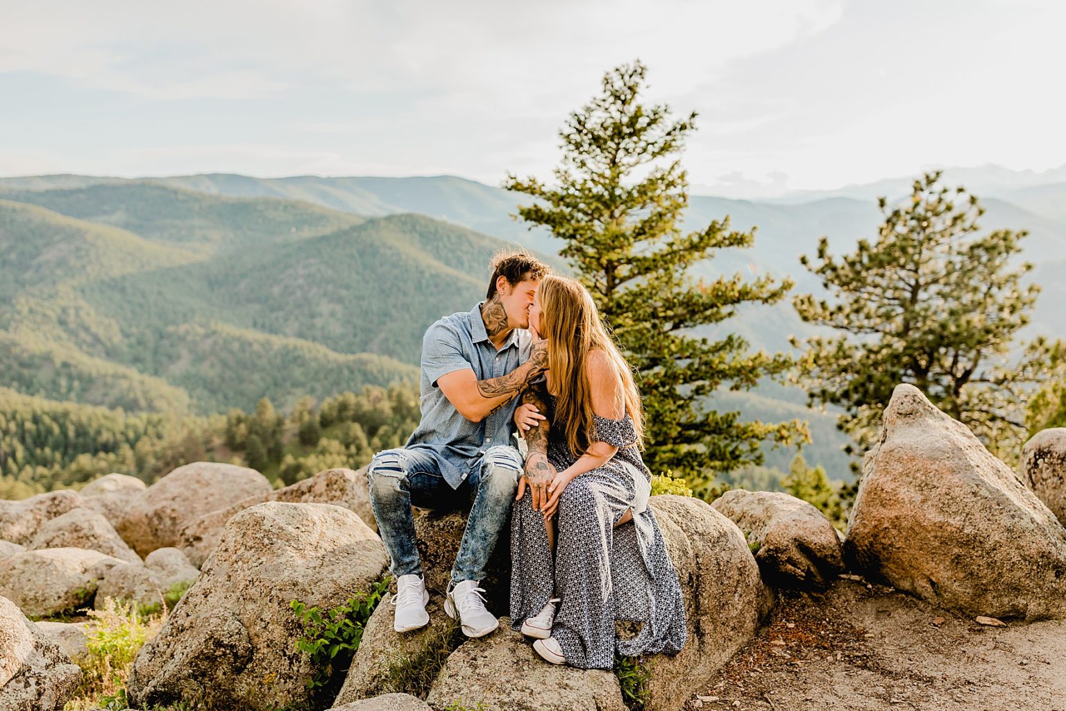 couple sits on a rock holding each other with green mountains and trees in the distance