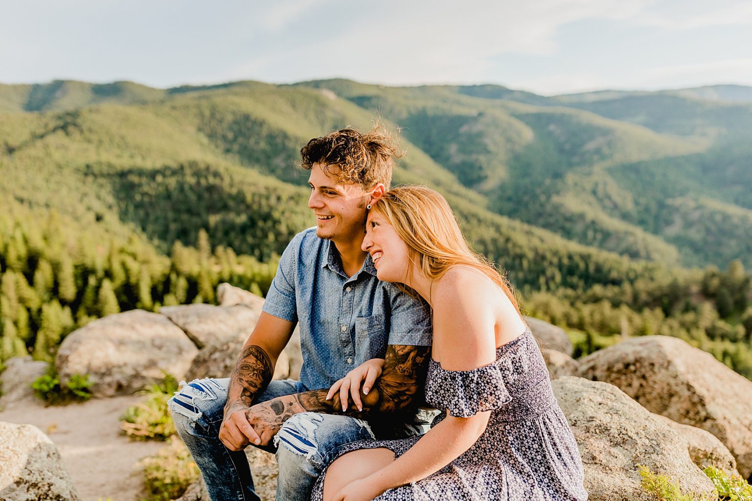 couple sits on a rock holding each other with green mountains and trees in the distance