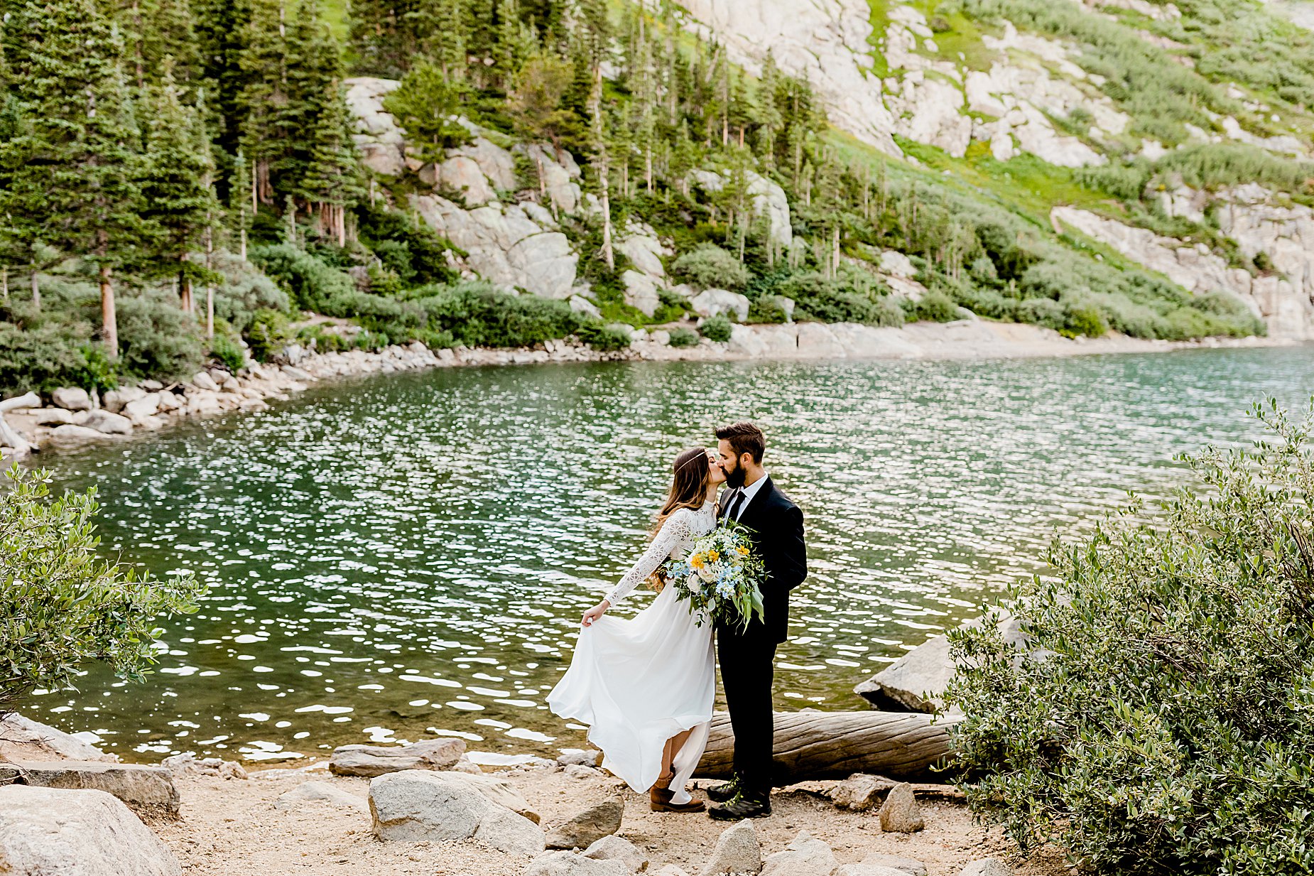 bride and groom stand in front of gorgeous blue alpine lake with evergreen trees in the background