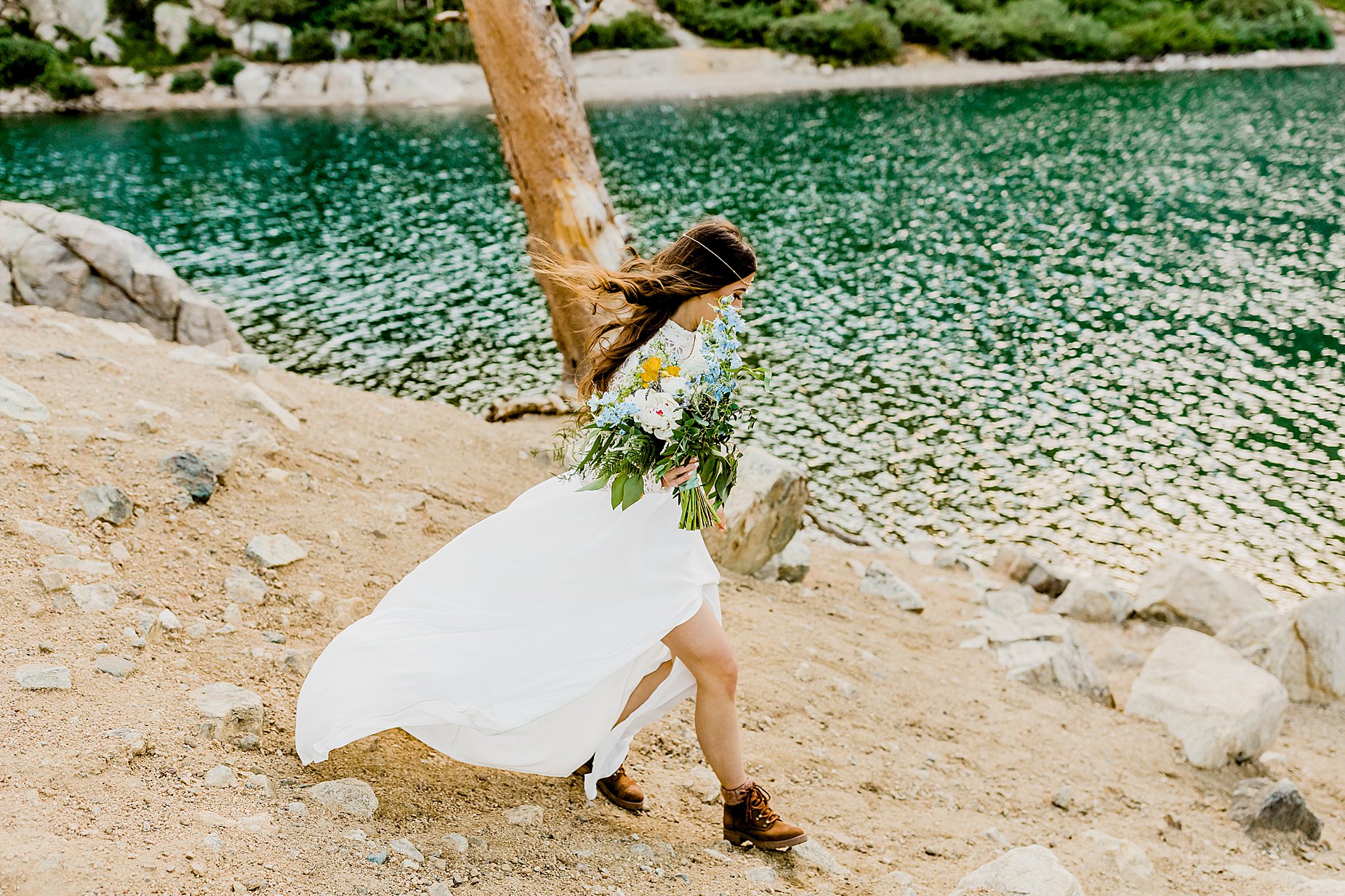 bride hikes down to gorgeous colorado lake in hiking boots while holding floral bouquet