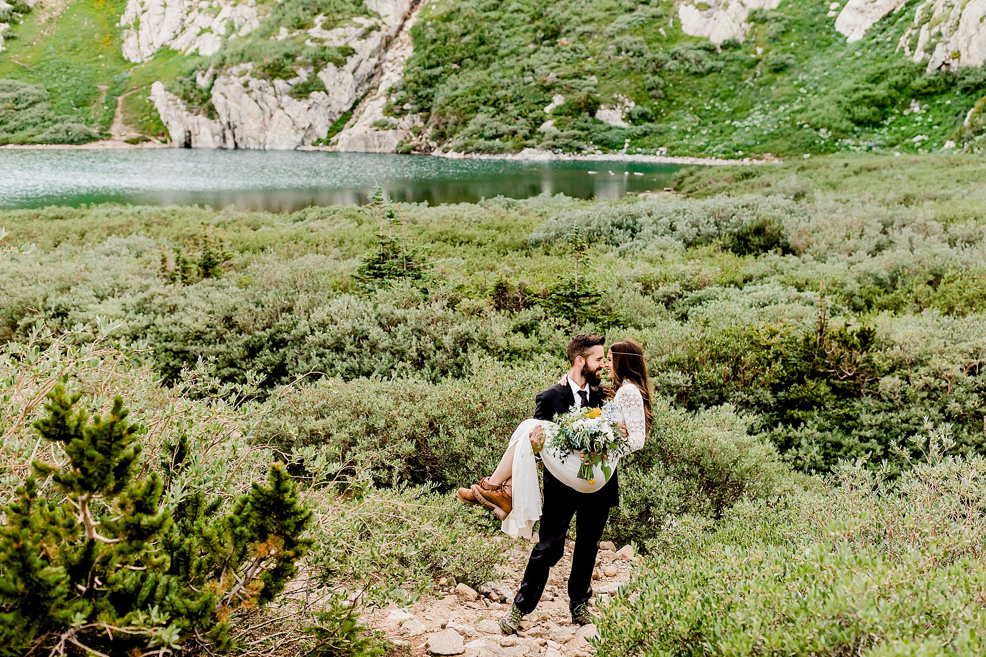groom holds bride in stunning colorado lake and mountain scenery / brides hiking boots are shown while she holds gorgeous bouquet