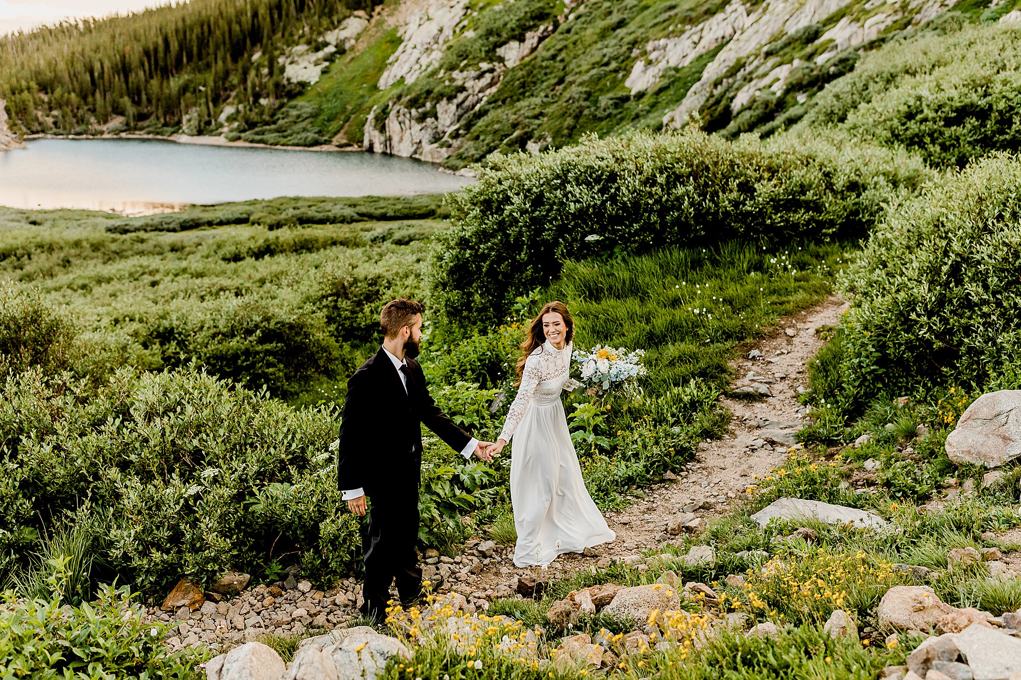 bride leads groom through stunning wildflowers in front of an alpine lake at st. mary's glacier for colorado elopement