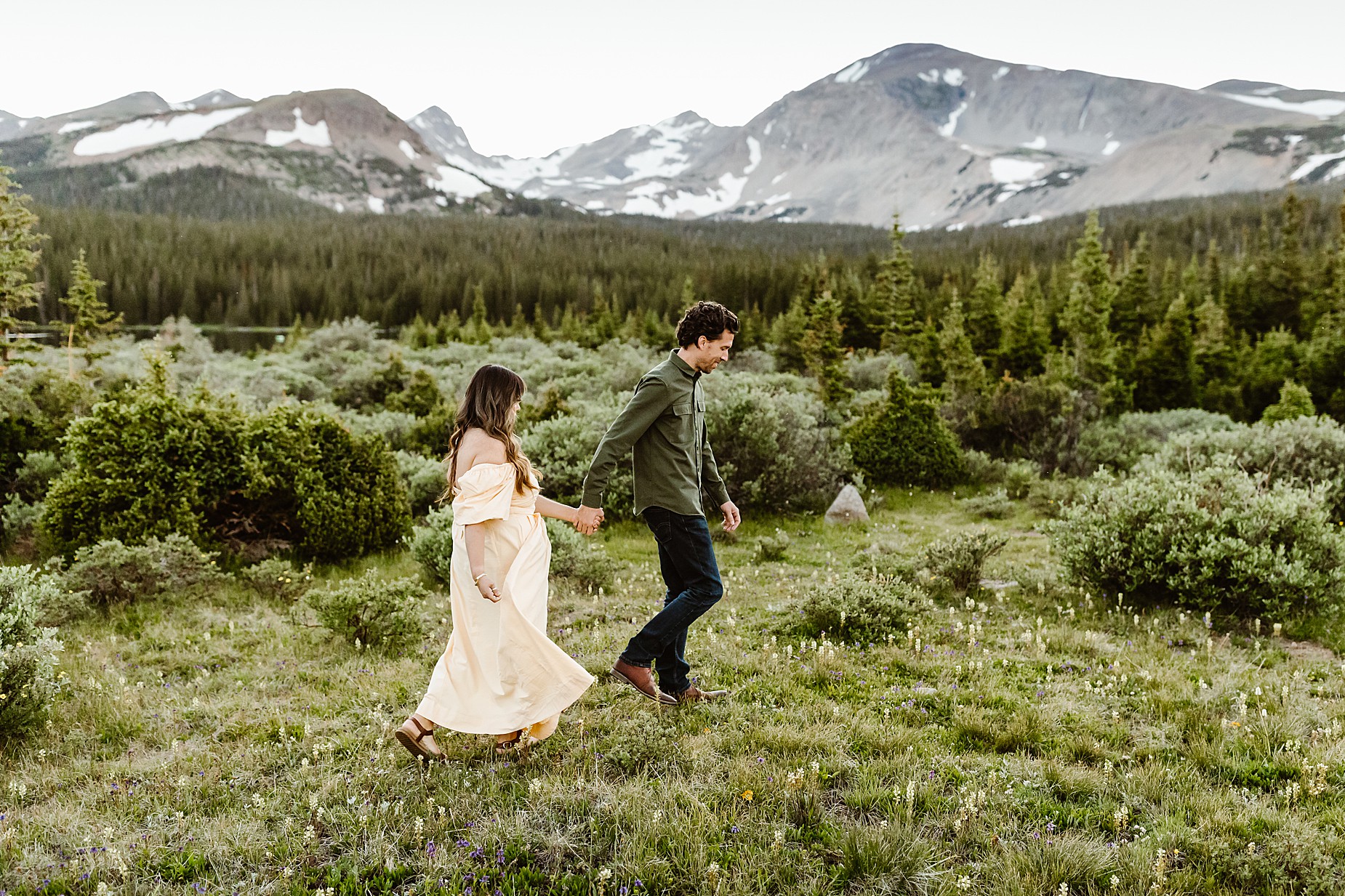 Couple takes anniversary photos in the Rocky Mountains in front of gorgeous alpine lake surrounded by greenery and wildflowers